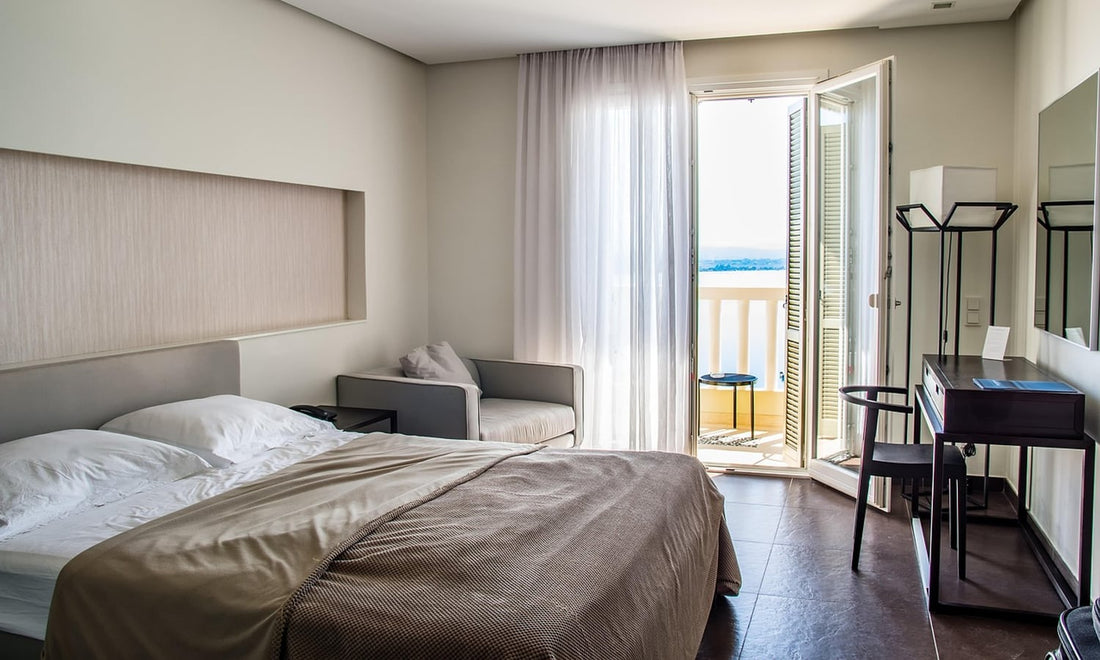 Bright hotel room with a made bed, an armchair, a small desk and an open balcony door
