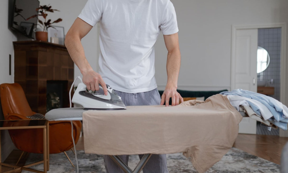 Man pressing a beige T-shirt with a traditional steam iron on an ironing board at home