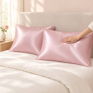 RosyIron blush pink satin pillowcases on two pillows on a made bed, a hand smoothing the satin