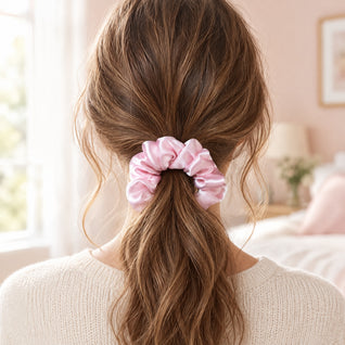 RosyIron pink scrunchie holding a woman's low loose ponytail, seen from behind in a bright bedroom